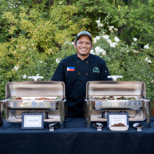 A man helping with catering from Malaya Eats. He is behind a table and in front of him are two catering dishes of food.