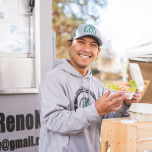 A man holding a to-go container of food and smiling. He is in a Malaya Eats hat and sweater.